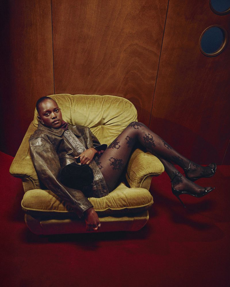 Woman with short hair poses in a yellow armchair indoors, wearing a metallic jacket, patterned tights, and high heels against a paneled backdrop.