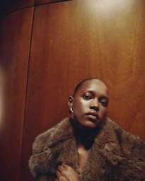 Model Alaato Jazyper posing in a plush brown fur coat against a warm wood-paneled wall for Mango.