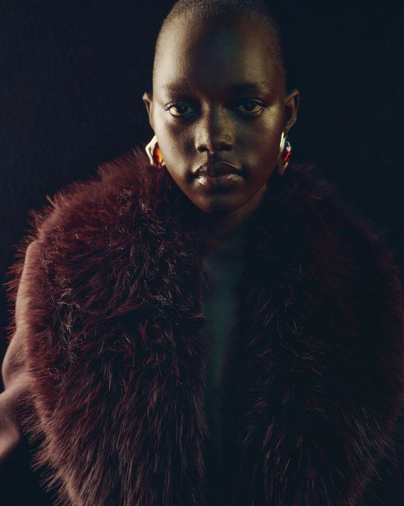 Model Alaato Jazyper wearing a burgundy faux fur collar and statement earrings against a dark seamless backdrop.