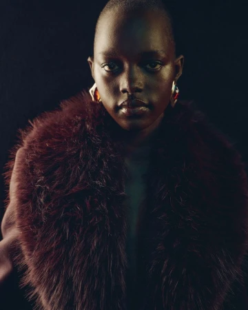 Model Alaato Jazyper wearing a burgundy faux fur collar and statement earrings against a dark seamless backdrop.
