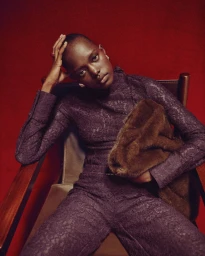 Model Alaato Jazyper seated in an armchair wearing a purple lace outfit and holding a brown fur clutch against a red backdrop.