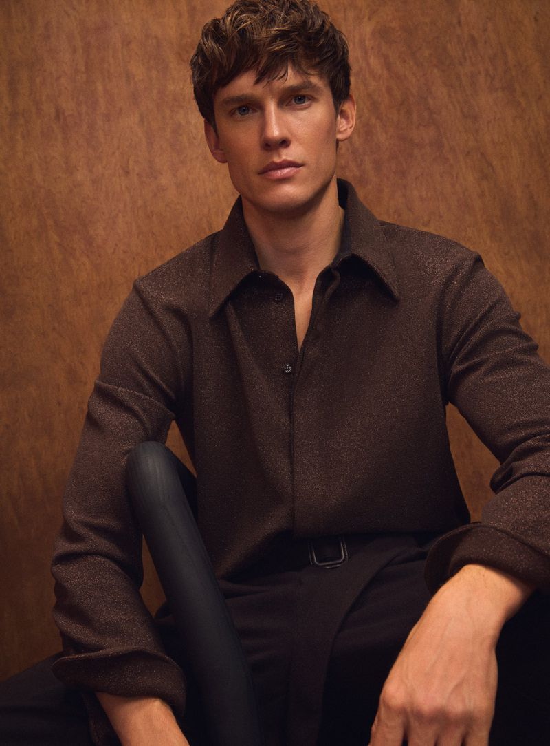 Portrait of a young adult male with brown hair wearing a brown collared shirt with black pants, posing in front of a brown background.