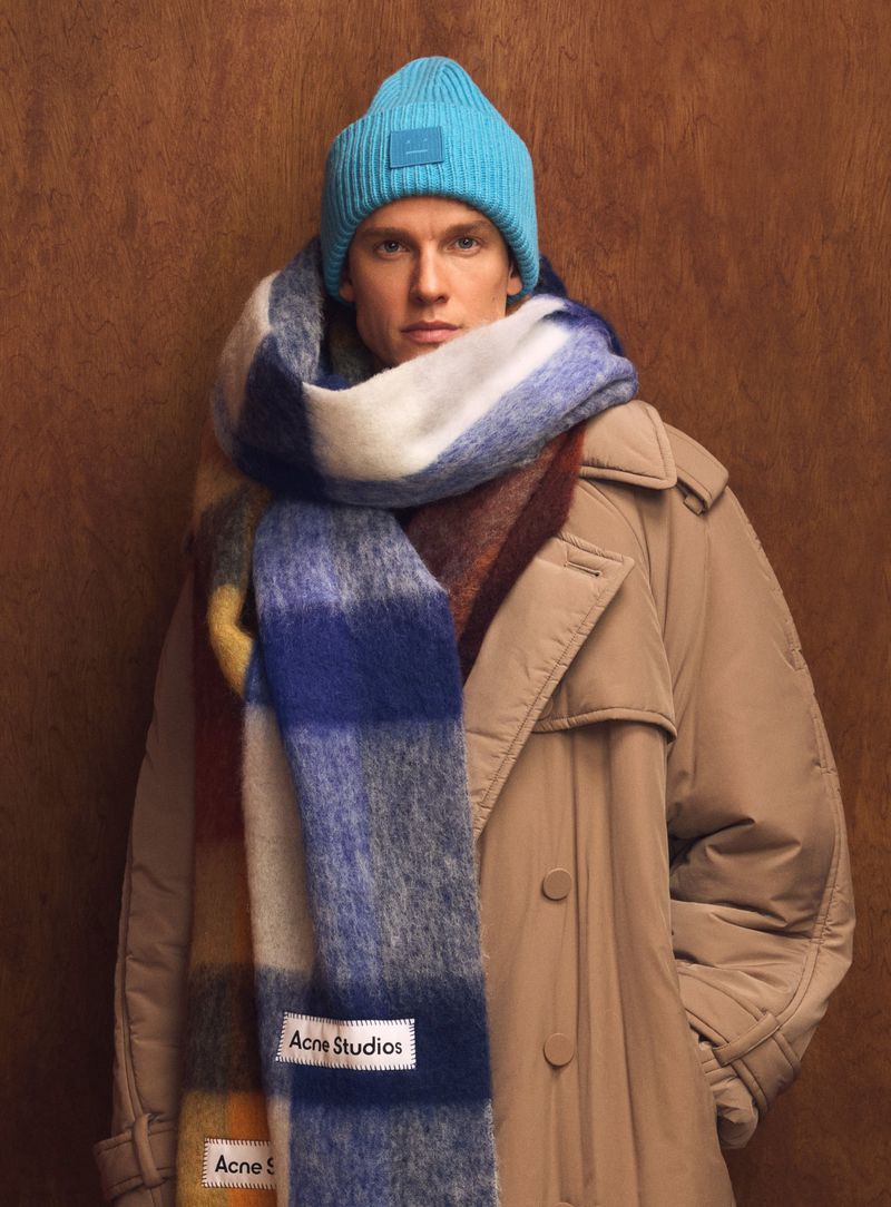 Male model in a tan trench coat, blue beanie, and oversized blue plaid scarf with the Acne Studios label against a brown background.
