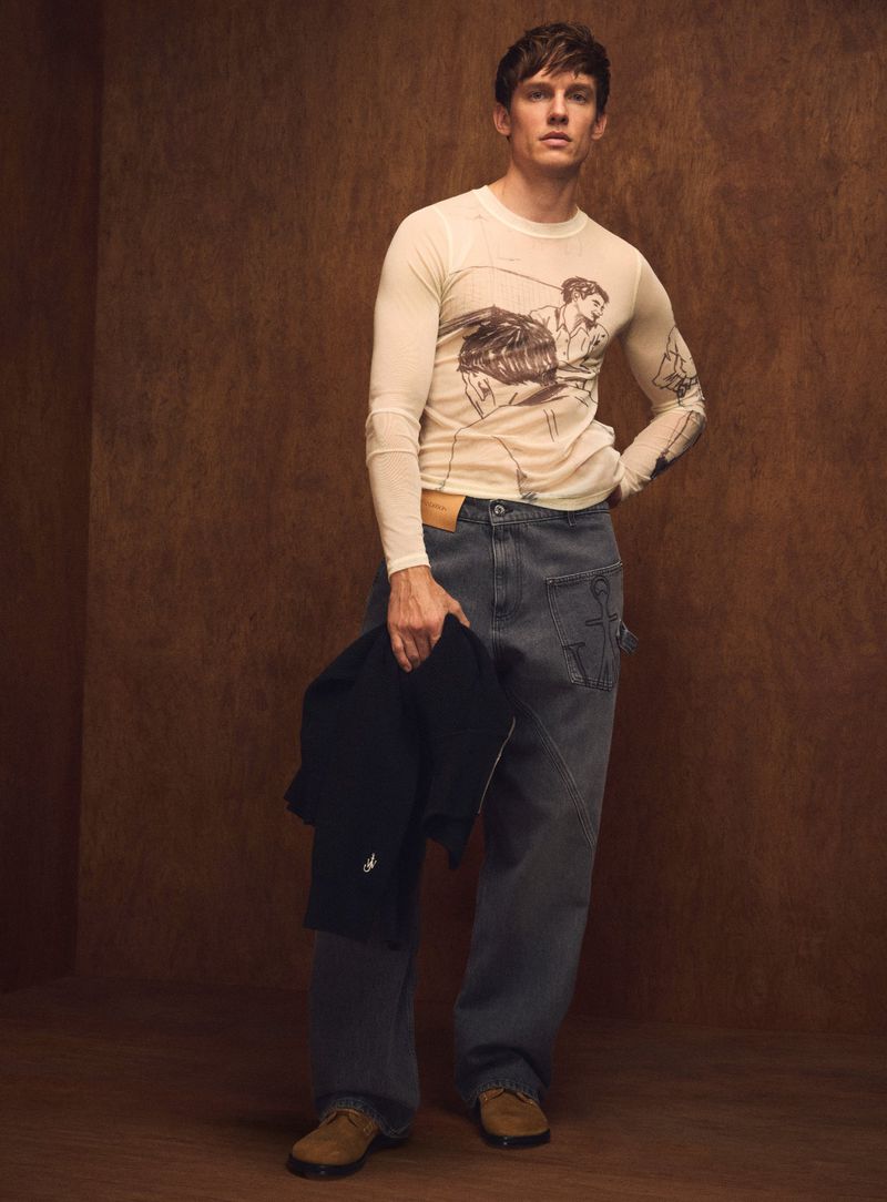 Full-length shot of a young adult male in casualwear with a graphic long-sleeve shirt, holding a dark jacket in a studio setting.