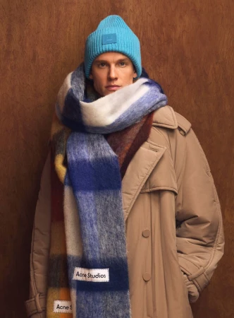 Model wearing a bright blue beanie and chunky plaid scarf wrapped around a beige coat against a dark wood background.