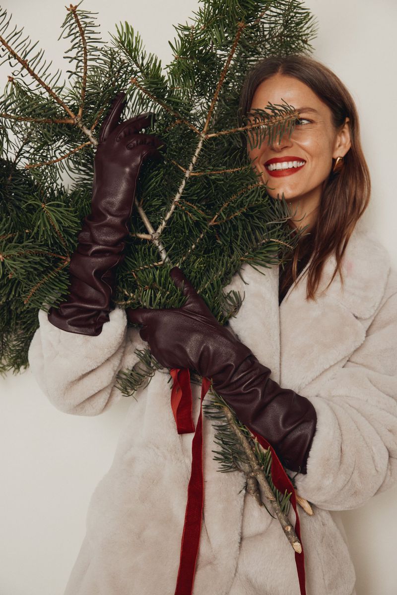 Smiling woman in a faux fur coat holding a branch of evergreen with red ribbons, wearing maroon leather gloves against white.