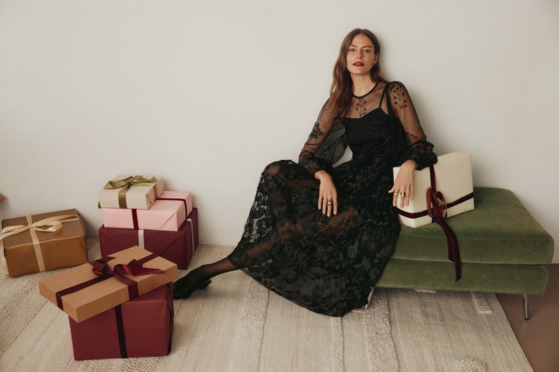 Elegant woman in a black lace dress sitting on a green ottoman in front of wrapped gift boxes. Fashion editorial shot, indoor setting.