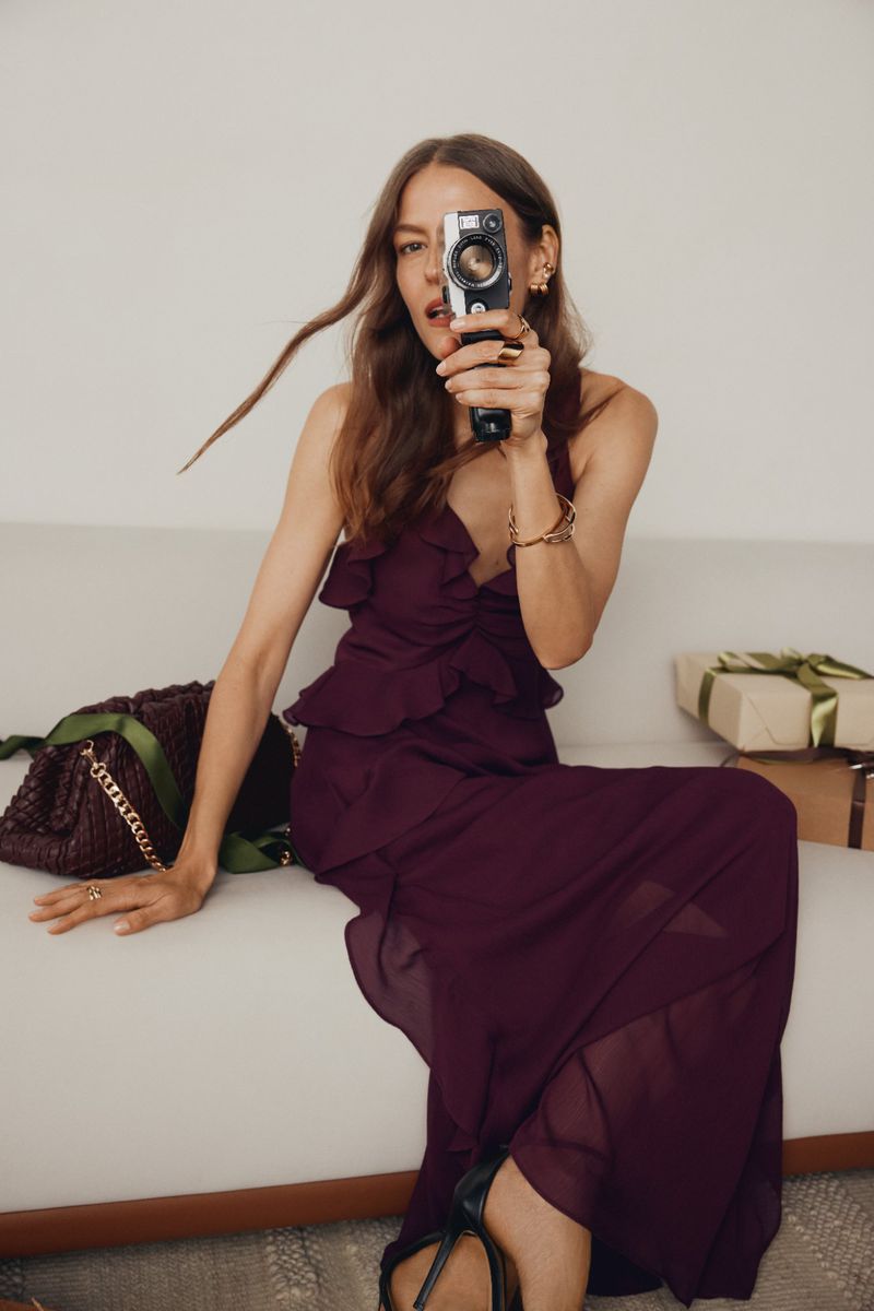 Woman with vintage camera in maroon ruffle dress on couch near wrapped gifts, embodying fashion, lifestyle, and potential celebration themes.