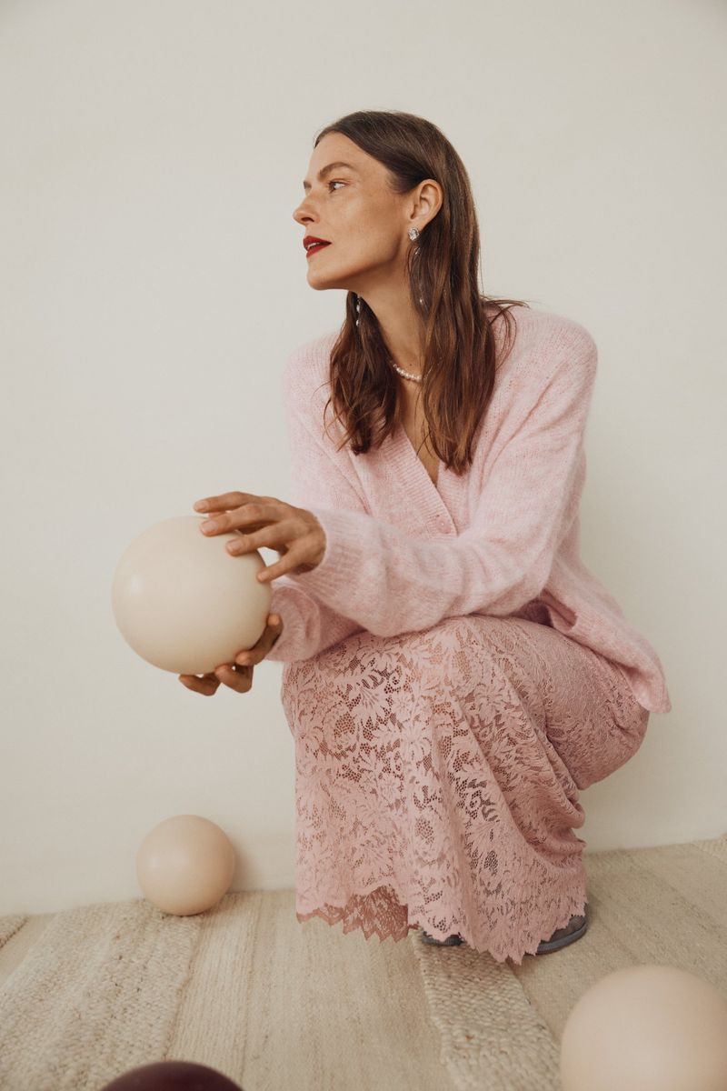 Woman in pink cardigan and lace skirt holding a balloon in studio setting, looking away from the camera with a neutral expression.
