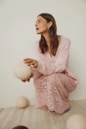 Viktorija Bauzyte crouching in a pink knit cardigan and floral lace skirt holding a spherical prop against a neutral wall.