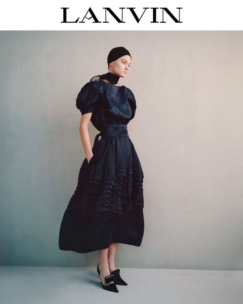 A woman models a navy blue gown with puffed sleeves and buckled heels against a neutral backdrop for a fashion editorial.