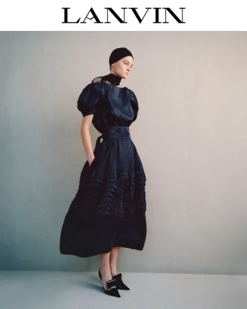 A woman models a navy blue gown with puffed sleeves and buckled heels against a neutral backdrop for a fashion editorial.