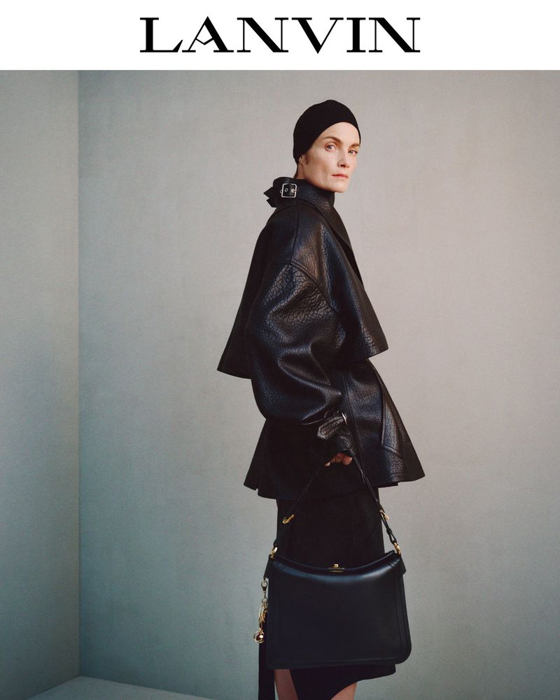 Model wearing a black leather jacket and cap holding a handbag against a plain grey studio wall.