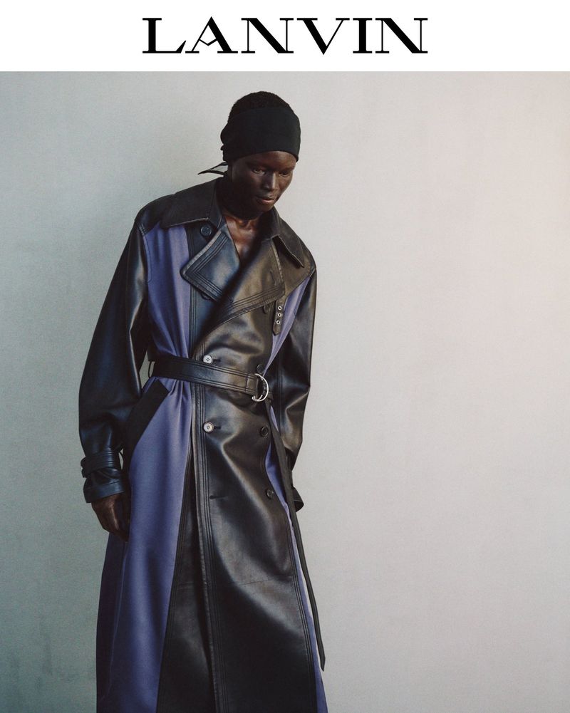 An adult fashion model wears a black and blue leather trench coat, head wrap, in a studio shot against a grey wall for Lanvin.