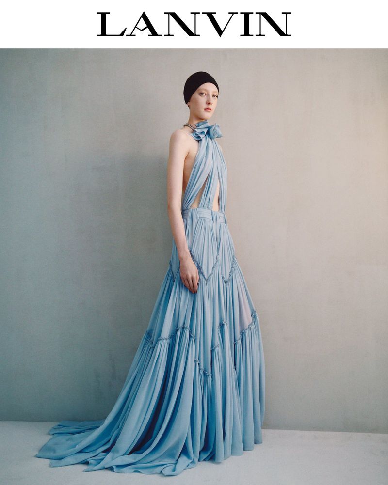 Elegant woman in light-blue Lanvin gown poses against textured wall, fashion editorial shot with studio lighting and soft color palette.