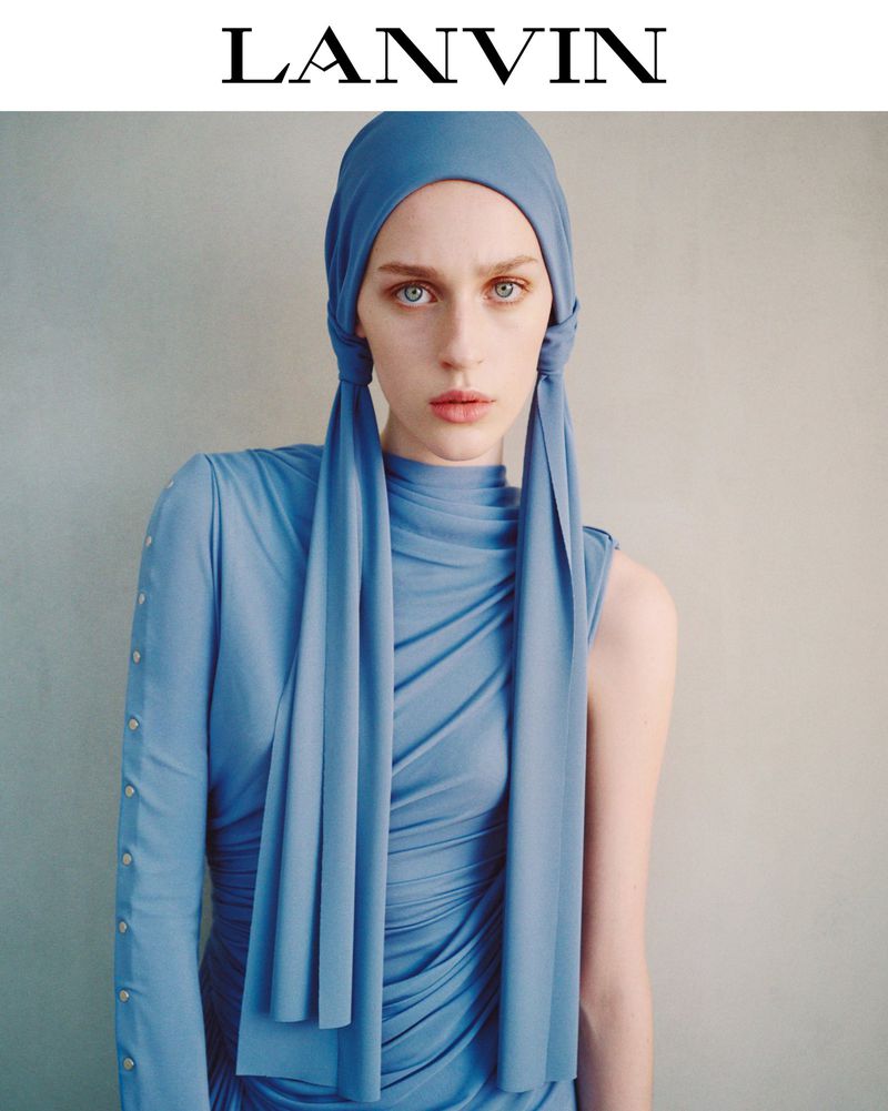 Fashion portrait of a fair-skinned woman wearing a blue headscarf and draped dress, staring directly at the camera with neutral expression.