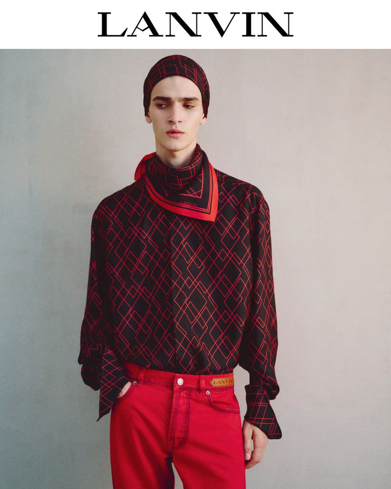 A slender adult male fashion model poses indoors wearing a black patterned shirt, red pants, and a matching red and black headscarf.