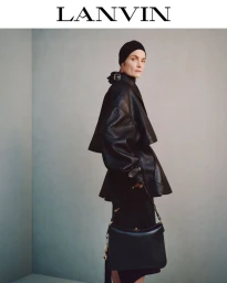 Model wearing a black leather jacket and cap holding a handbag against a plain grey studio wall.