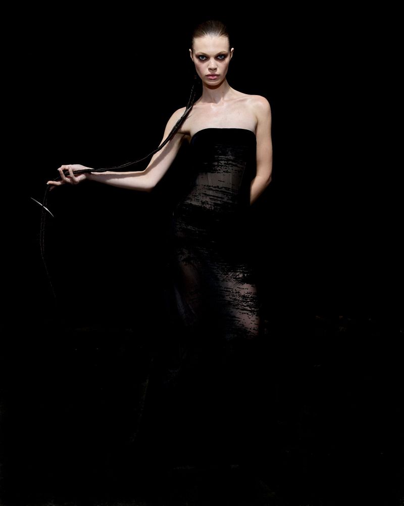 Fashion shot of a model in a black strapless dress holding a long black extension against a dark background in a studio setting.