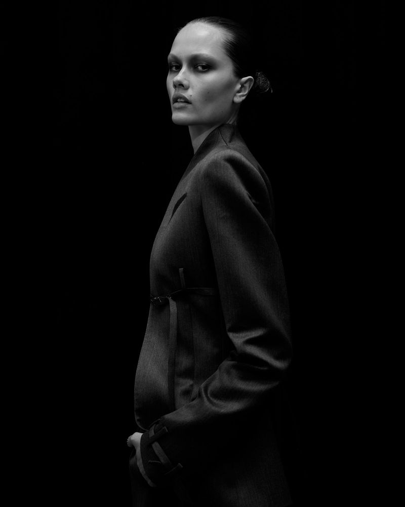 Model wearing a dark tailored blazer in profile against a black studio background in a black-and-white fashion portrait.