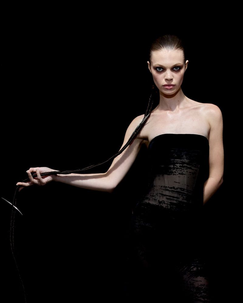 Fashion shot of a slender adult female model in a black strapless dress, holding a black braid against a dark studio backdrop.