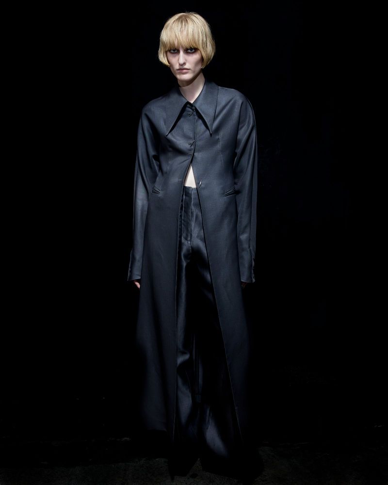 Person in a long dark coat and matching pants standing in front of a black backdrop in a studio setting. Fashionable attire and pose.