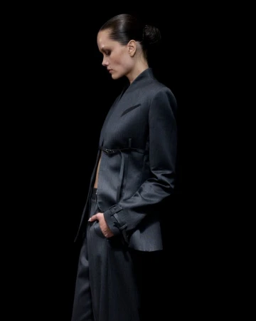 Model in profile wearing a tailored dark grey belted blazer and matching trousers against a solid black background.