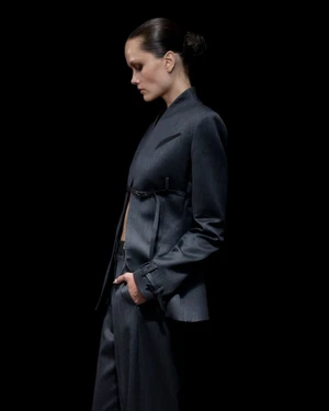 Model in profile wearing a tailored dark grey belted blazer and matching trousers against a solid black background.