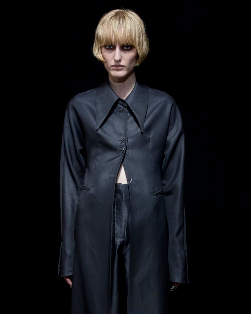 Model with blonde bob wearing a dark buttoned coat against a black background for Grace Ling S/S 26