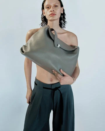 Model holds a large grey leather handbag. The model wears wide, high-waisted grey pants with a black tie belt, against a white backdrop.