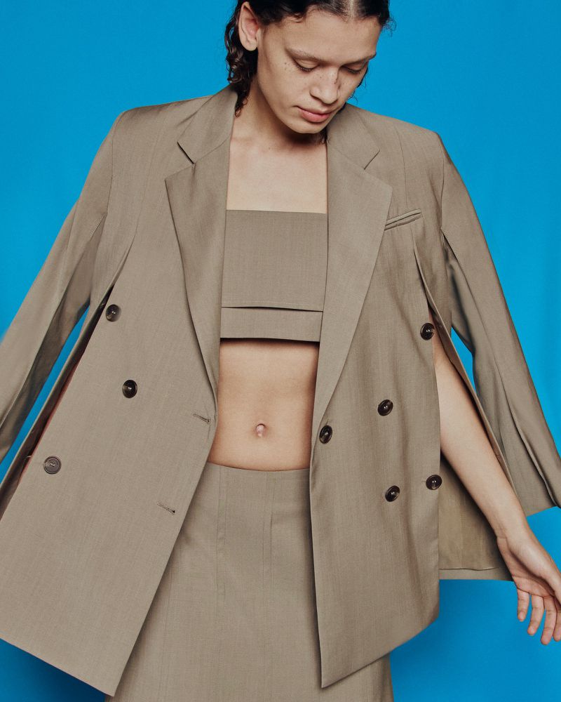Adult female in a studio-lit fashion shot wearing a tan blazer with cutout sleeves, skirt, and matching crop top against a blue background.