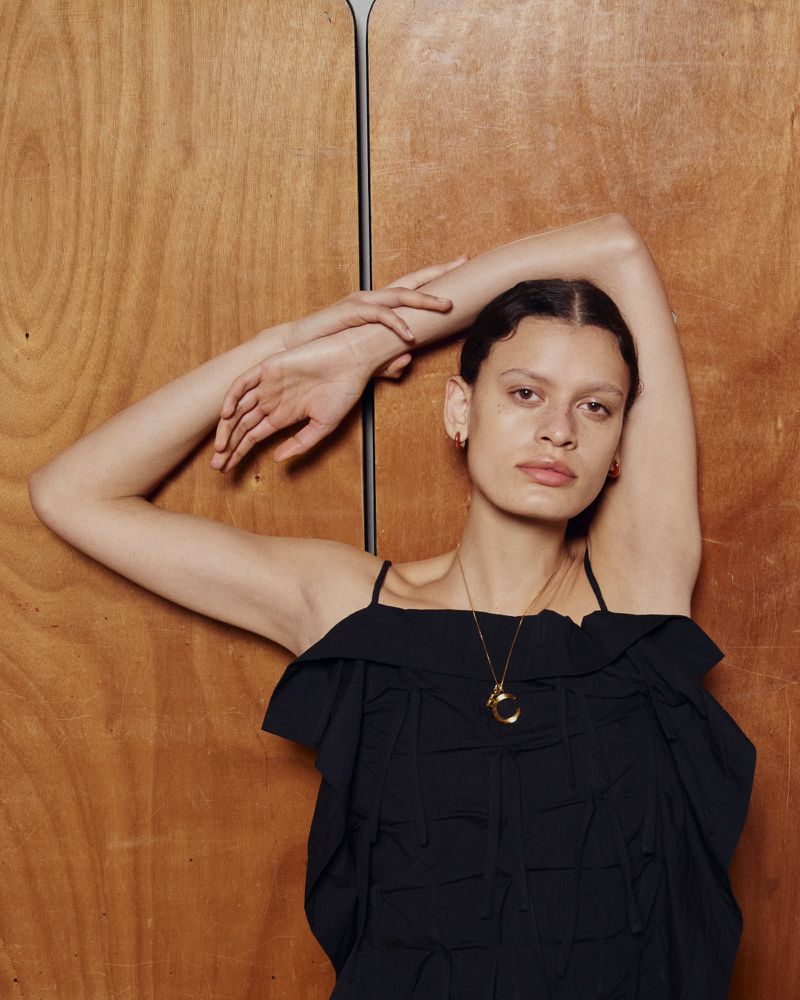Model posing against a wood paneled wall wearing a black ruffle top and gold necklace for Eudon Choi SS24 campaign