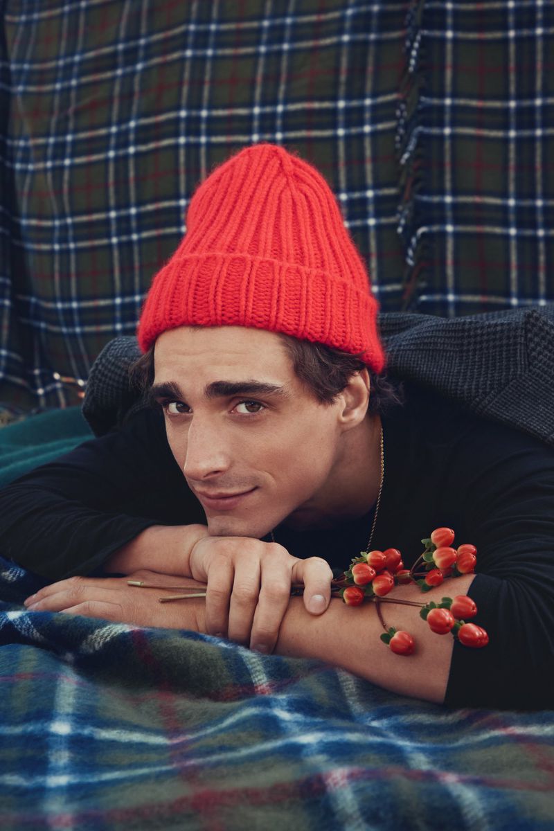 Portrait of a young adult male with a red knit beanie, lying down, holding red berries with a plaid background. Dark hair, looking at camera.