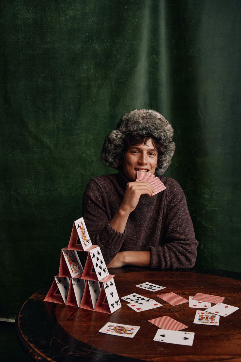 Portrait of a young adult in a furry hat, biting a playing card, with a card pyramid on a wooden table against a green backdrop.