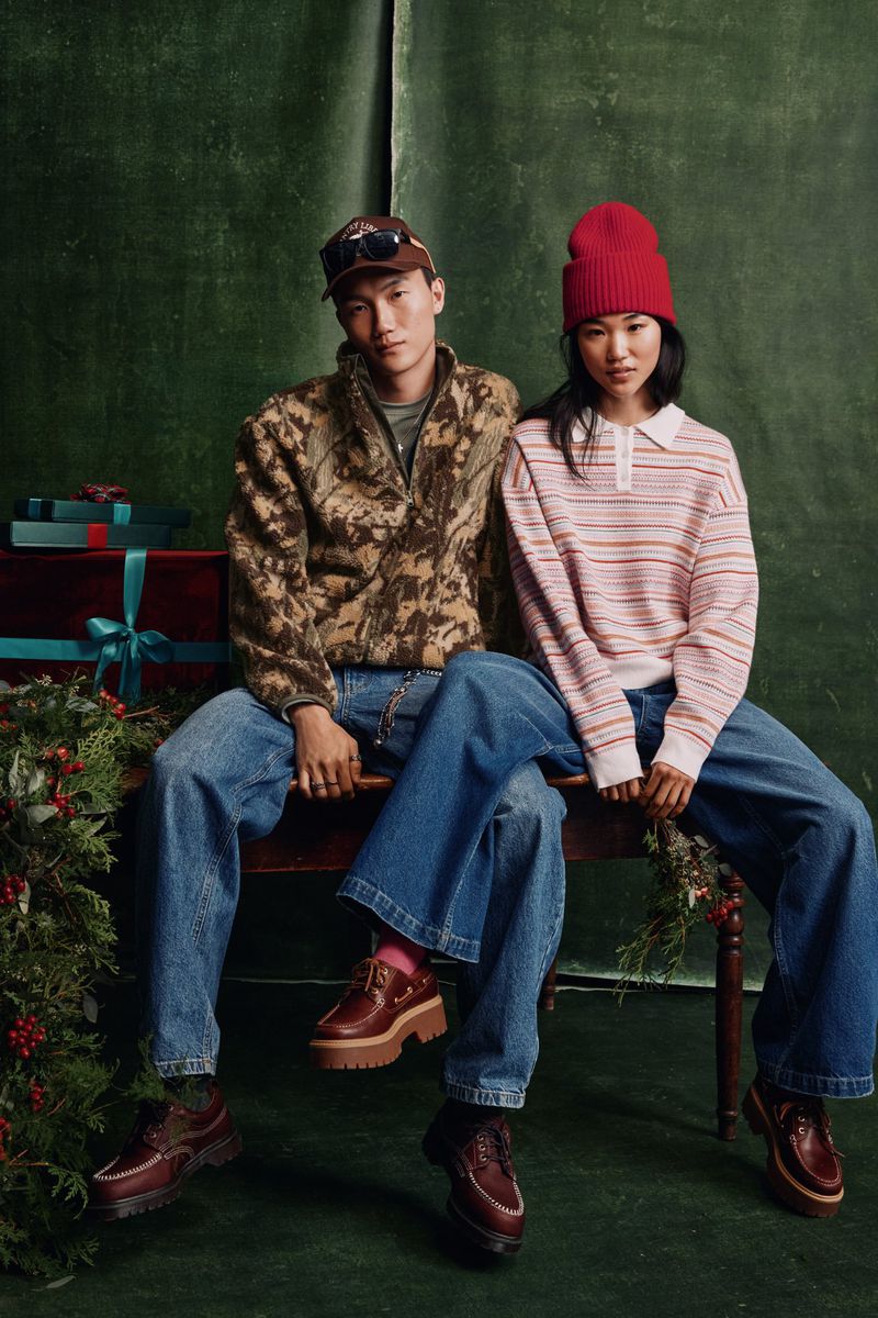 A stylish Asian man and woman sit together wearing casual fall or winter clothing with presents and greenery in a warm, inviting scene.