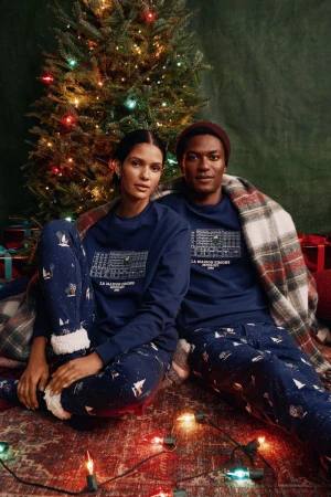 A stylish mixed-race couple in matching pajamas poses indoors with a Christmas tree and presents, creating a festive, cozy holiday scene.