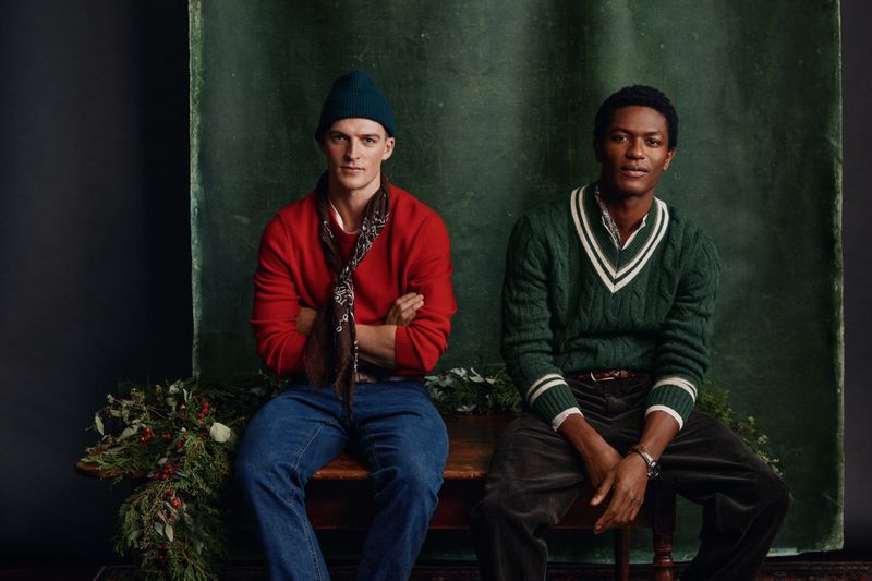 Two men pose indoors in sweaters and hats. One man wears red, the other green. Winter greenery is arranged on the surface below.