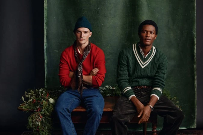 Two men pose indoors in sweaters and hats. One man wears red, the other green. Winter greenery is arranged on the surface below.