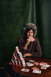 Man wearing a fur ushanka hat and brown sweater building a card house on a round wooden table