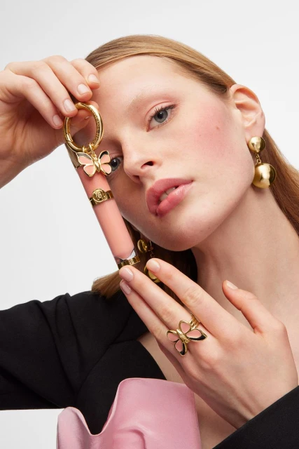 Close-up of a woman holding lipstick and wearing jewelry. A pink ruffled fabric accentuates her fashionable attire in a studio setting.