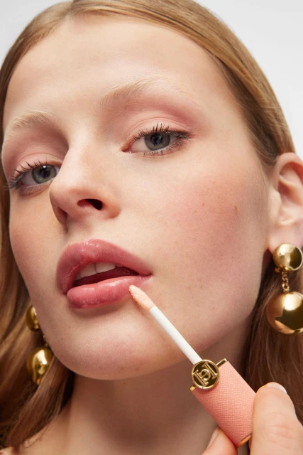 Close-up of an adult female applying lip gloss with a wand applicator. She has fair skin, light hair, and is wearing gold earrings.