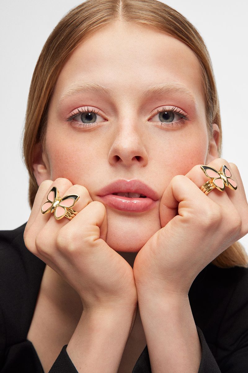Close-up portrait of a blonde woman with two hands framing face, wearing butterfly rings on fingers. Soft lighting, pale pink makeup.