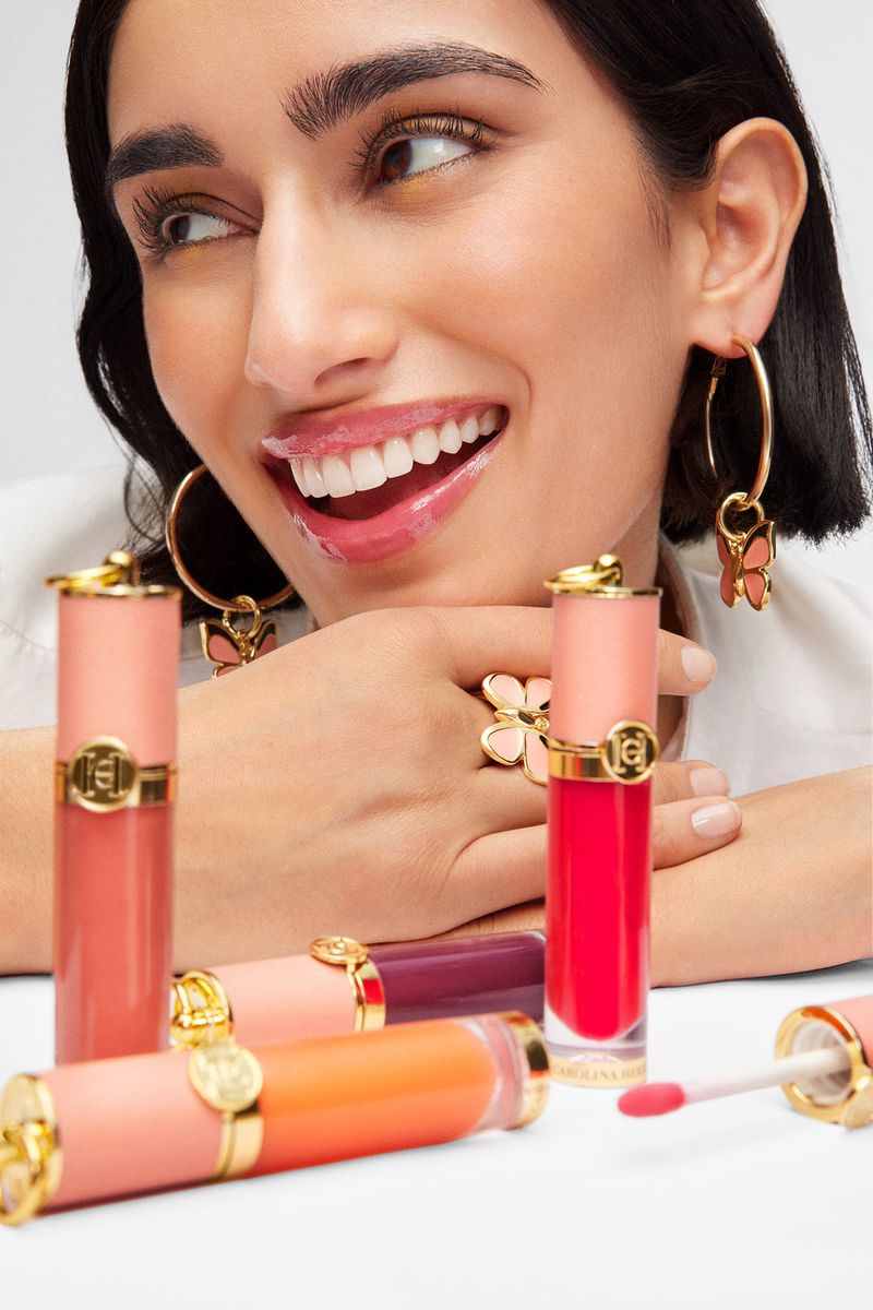 Close-up of a smiling woman with jewelry posing with colorful lipsticks on a white surface in a beauty and fashion product shot.