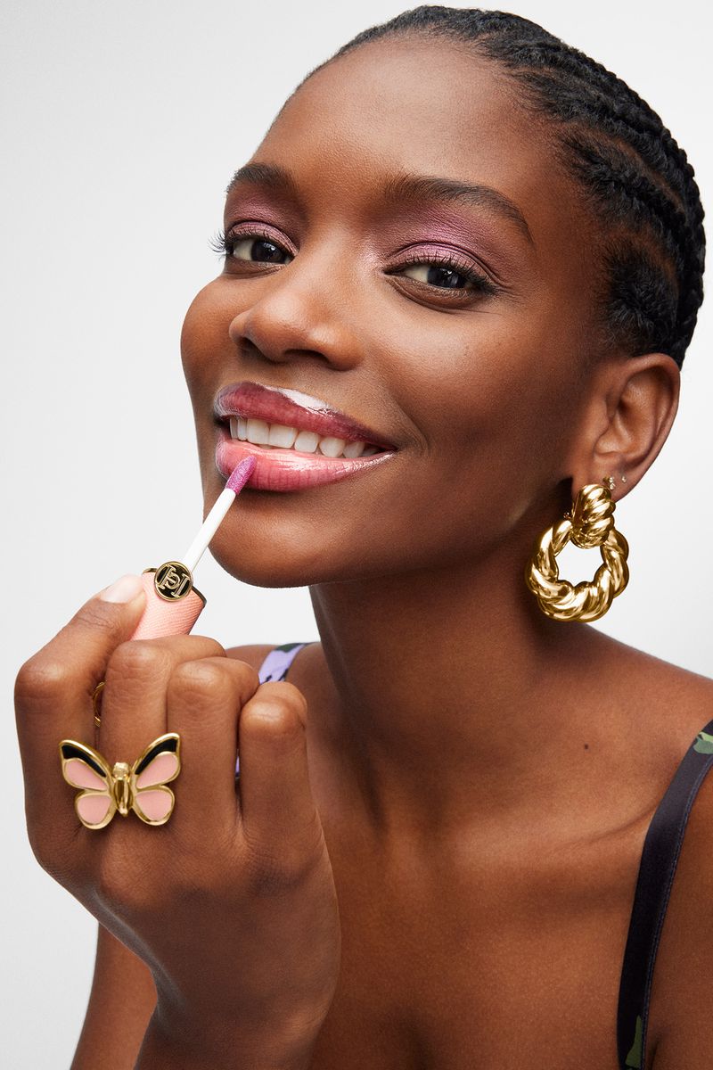 Close-up of a Black woman applying lip gloss; she is wearing large gold earrings and a butterfly ring against a white backdrop.