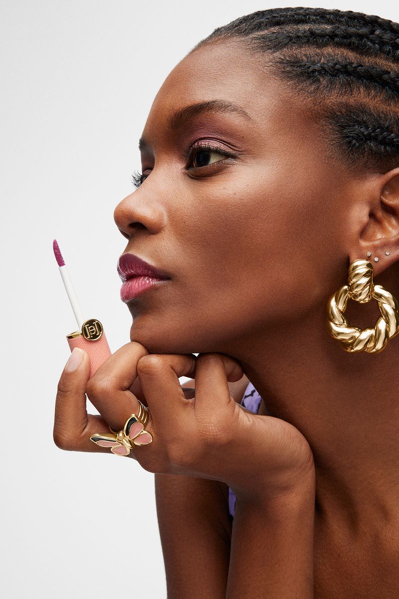 Beauty shot of an African-descent woman with plum lipstick and gold jewelry holding a lip gloss wand against a white backdrop.