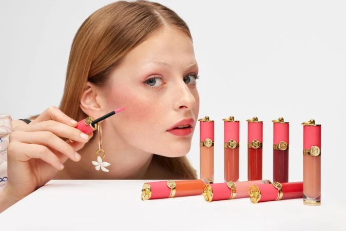 Fair-skinned model applies pink lip gloss, surrounded by tubes of various shades, on a white table against a bright white backdrop.