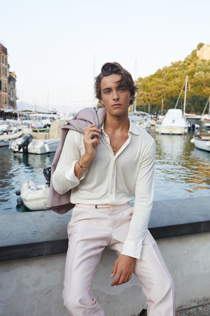 Handsome man with auburn hair poses outdoors by a harbor wearing a white shirt, light pink pants, and a jacket over his shoulder.