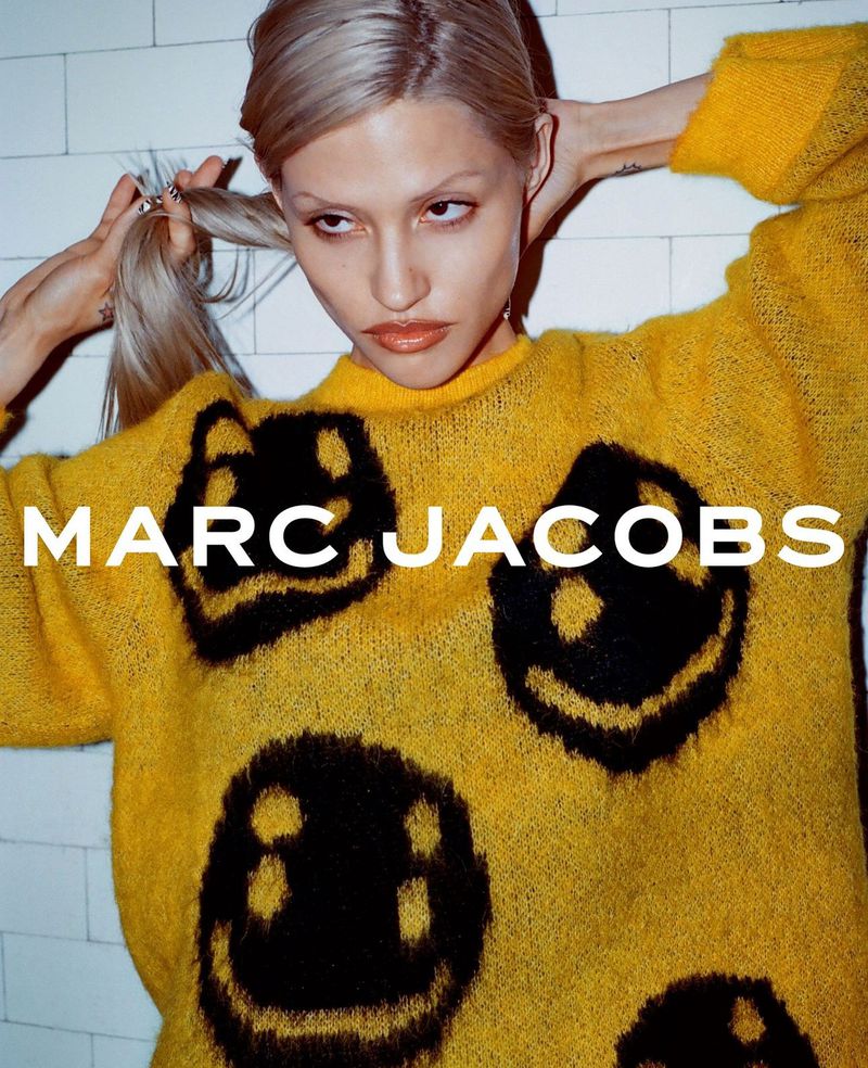 A fair-skinned adult woman with blonde hair, wearing a yellow sweater with black graphic smiley faces, and the brand name Marc Jacobs.