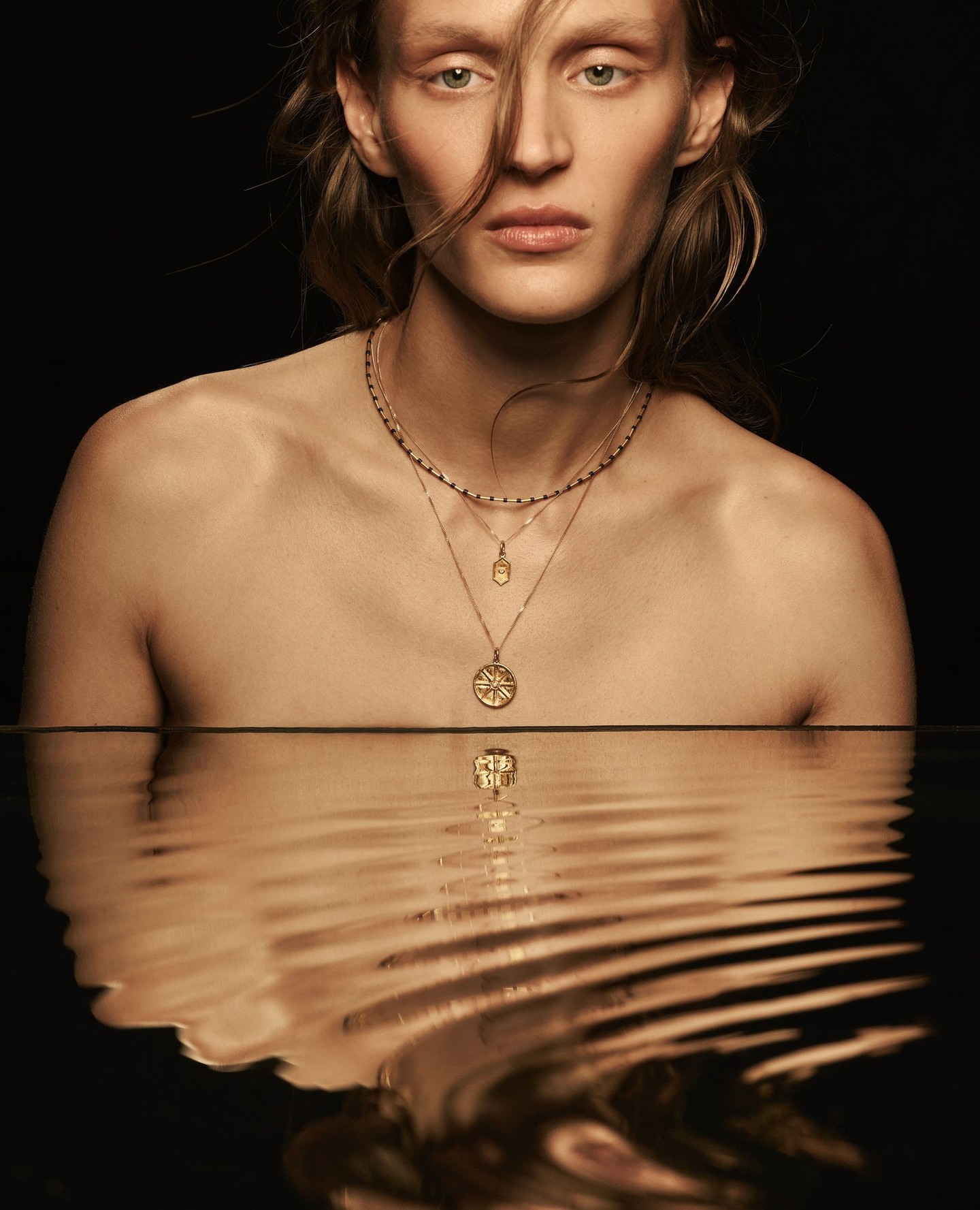 Elegant studio shot of a fair-skinned adult female model wearing gold necklaces, reflected in a sleek water surface against a dark backdrop.
