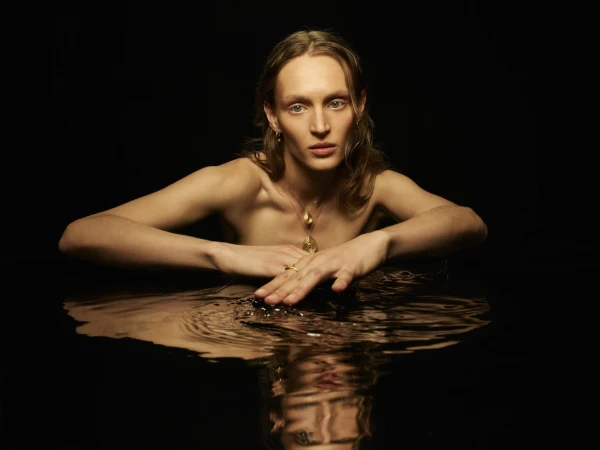 Model wearing gold jewellery leaning over dark reflective water for Temple of the Sun Reflections campaign